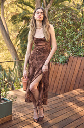 A young woman is standing outdoors on a wooden deck. She is wearing a sleeveless, brown dress with a floral pattern. The dress has a high slit on one side, revealing her leg. She has on high-heeled shoes and accessories including hoop earrings, a necklace, a bracelet, and a ring.